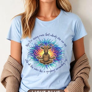 Op de afbeelding: Lichtblauw T-shirt met een mediterende geit omringd door een kleurrijk, wervelend ontwerp. De tekst luidt: "Nu zie ik muziek die niemand anders kan zien met alle kleuren als een symfonie om me heen." Het shirt is gelabeld "LICHTBLAUW".