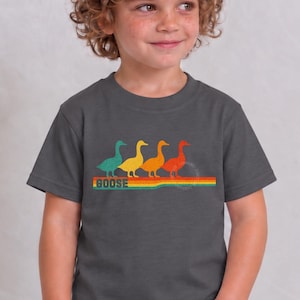 May include: A dark gray short-sleeve t-shirt featuring a graphic of four ducks in teal, yellow, orange, and red, walking in a line above a multi-colored horizontal striped bar with the word GOOSE printed in a bold, retro-style font underneath.