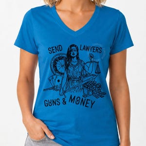 May include: A bright blue V-neck t-shirt featuring a black graphic print of Lady Justice holding scales and a sword. The design includes a revolver, playing cards, a chest of money, and the text SEND LAWYERS GUNS & MONEY in black ink.