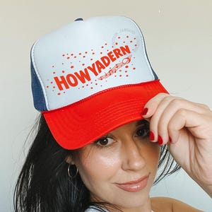 Puede incluir: Gorra de camionero roja, blanca y azul con el texto "HOWYADERN" en letras rojas. La gorra tiene un panel frontal blanco, una visera roja y una parte trasera de malla azul. La gorra se está usando.