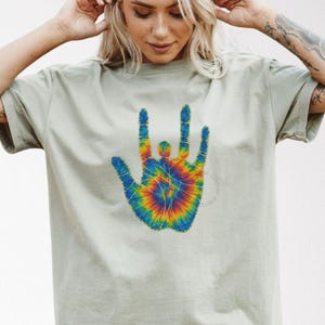 Könnte beinhalten: Ein hellgrünes T-Shirt mit einem Batik-Hand-Symbol in leuchtenden Farben. Das Handsymbol ist in Blau-, Gelb-, Orange- und Grüntönen gehalten. Das Shirt hat kurze Ärmel und eine lockere Passform.