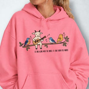 May include: A bright pink hooded sweatshirt featuring a graphic of a white goat playing an acoustic guitar while perched on a branch with four colorful birds. The text below reads, I'D SING ALONG WITH THE BIRDS, IF I ONLY KNEW THE WORDS.