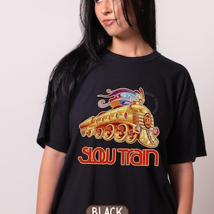 Puede incluir: Camiseta negra con una ilustración dorada y roja de un tren y el texto "SLOW TRAIN" en rojo. El diseño del tren incluye detalles coloridos y el número 33. La palabra "BLACK" está impresa en la parte inferior.