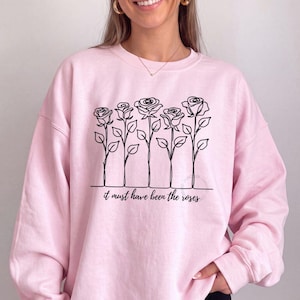 May include: A light pink crewneck sweatshirt featuring a black line art illustration of five roses on stems. Below the floral design, the text "it must have been the roses" is printed in a black cursive script font across the center of the chest.