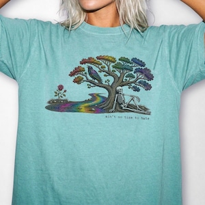 May include: A teal short-sleeve t-shirt featuring a graphic of a skeleton sitting under a rainbow-colored tree with a bird, a rainbow stream, and a rose. The text "ain't no time to hate" is printed in black below the graphic illustration.