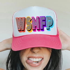 May include: A pink and white trucker hat with the text "WSMFP" in colorful, layered lettering. The hat has a pink brim and mesh backing. The wearer is smiling and sticking out their tongue.