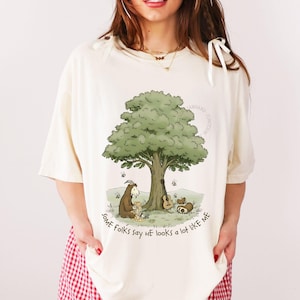 May include: Cream-coloured t-shirt with a cartoon illustration of a tree, a goat-like creature, and a guitar. The text "SOME Folks say HE looks a lot like ME" is printed on the shirt. The shirt has short sleeves.