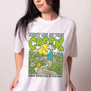 May include: White t-shirt with a colorful graphic featuring a goat fishing in a creek. The text "MEET ME AT THE CREEK" and "MUDDY WATER TAKE MY PAIN AWAY" are printed above and below the illustration.