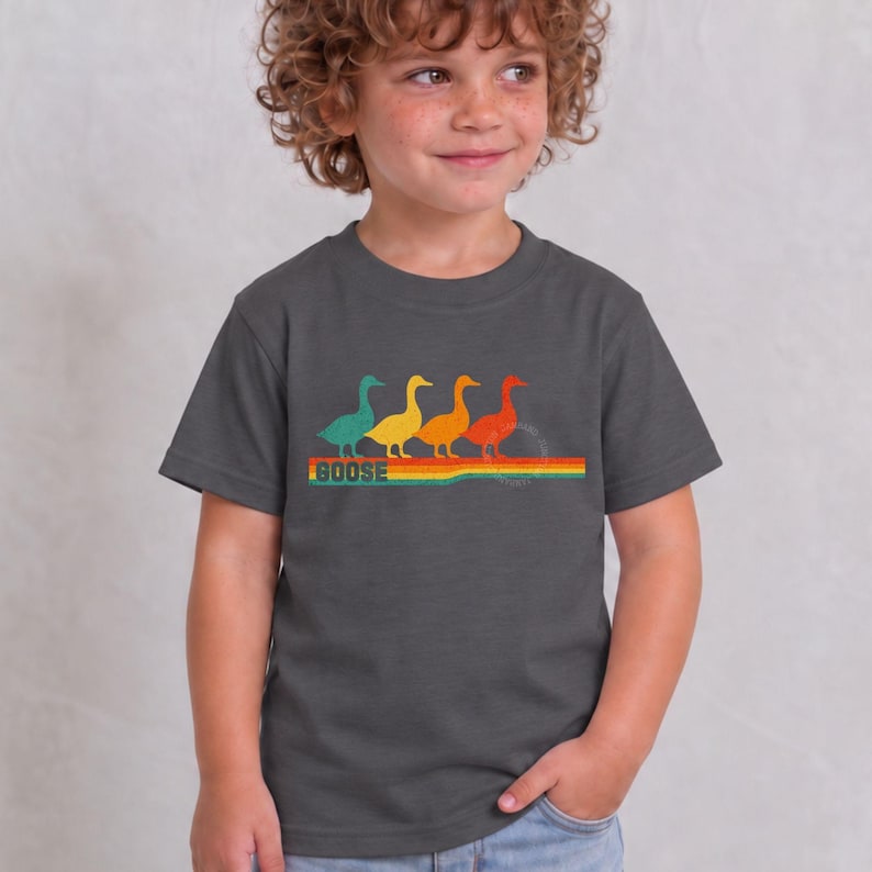 May include: A dark gray short-sleeve t-shirt featuring a graphic of four ducks in teal, yellow, orange, and red, walking in a line above a multi-colored horizontal striped bar with the word GOOSE printed in a bold, retro-style font underneath.