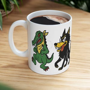 May include: White ceramic mug with a handle, filled with dark coffee. The mug features cartoon illustrations of a green alligator with a yellow bandana and a black wolf with a red tongue. The alligator and wolf are holding forks.