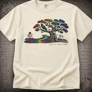 May include: A cream-colored t-shirt featuring a graphic of a skeleton resting under a rainbow-colored tree with a bird perched on a branch. A rainbow stream flows nearby, and a single rose stands alone. The text reads, "ain't no time to hate."
