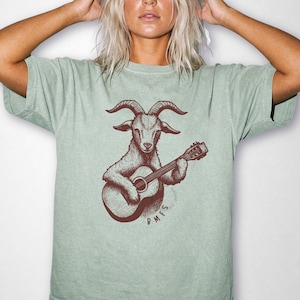 Può includere: Una t-shirt a maniche corte color verde salvia con un'illustrazione grafica marrone scuro di una capra che suona una chitarra acustica. Il testo BMFs è stampato in lettere marrone scuro sotto l'illustrazione della capra sul davanti.