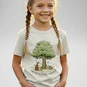 Puede incluir: Camiseta beige claro con una ilustración caprichosa de un árbol con un personaje parecido a una cabra, una guitarra y un oso. El texto "SOME FOLKS say HE looks a lot LIKE ME" está impreso en un círculo alrededor de la base del árbol.