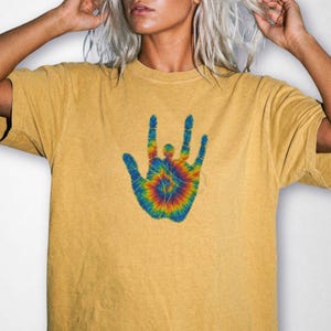May include: A mustard yellow short-sleeve t-shirt featuring a central graphic of an open hand in a tie-dye pattern. The hand design displays a vibrant swirl of blue, green, yellow, orange, and red colors, with fingers spread in a gesture.
