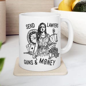 May include: White ceramic mug featuring a black illustration of Lady Justice with scales and a sword, alongside a roulette wheel, playing cards, a gun, and money. The text on the mug reads "SEND LAWYERS GUNS & MONEY."