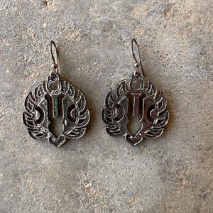 May include: A pair of silver-toned drop earrings. Each earring features a decorative, openwork design with a central vertical element and surrounding ornate details. The earrings have a hook closure.