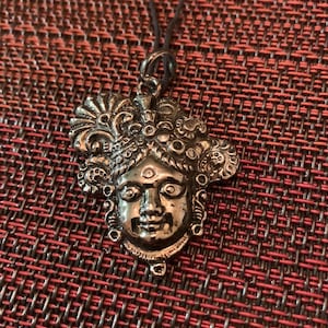 May include: Silver-toned pendant featuring a detailed face with ornate headdress and facial features. The pendant is suspended from a black cord. The design has a vintage or antique aesthetic, with intricate detailing.