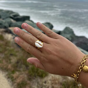 May include: A gold-toned ring featuring a white cowrie shell centerpiece, worn on a finger. The ring has a smooth, polished metal band that contrasts with the natural texture of the shell. A gold-toned chain bracelet with small charms is also visible.