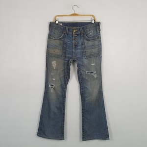 May include: Light blue denim jeans with a button fly and distressed details. The jeans have a flared leg, multiple pockets, and a worn appearance. The jeans are hanging on a wooden hanger.