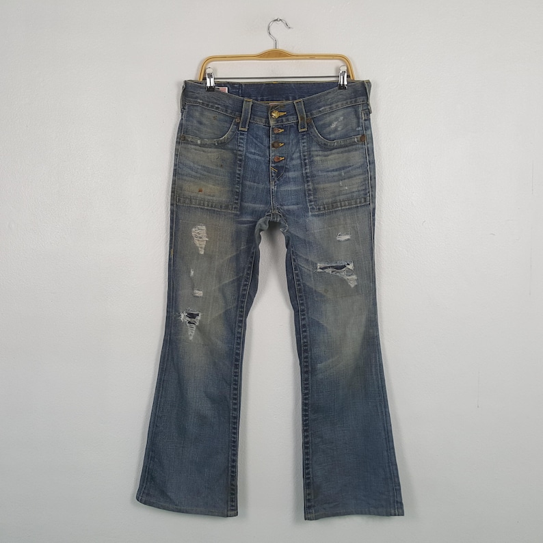 May include: Light blue denim jeans with a button fly and distressed details. The jeans have a flared leg, multiple pockets, and a worn appearance. The jeans are hanging on a wooden hanger.