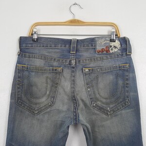 May include: Faded blue denim jeans with a distressed look, featuring a classic five-pocket design. The back pockets have a signature curved stitch. A leather patch with a skull and rose design is visible on the waistband.