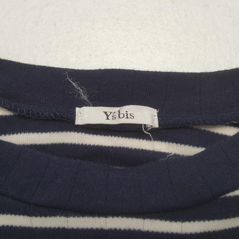 May include: Close-up of a navy blue and white striped top. The top has a ribbed texture and a rounded neckline. A small tag with the text "Y's bis" is sewn into the neckline. The fabric appears to be a soft knit.