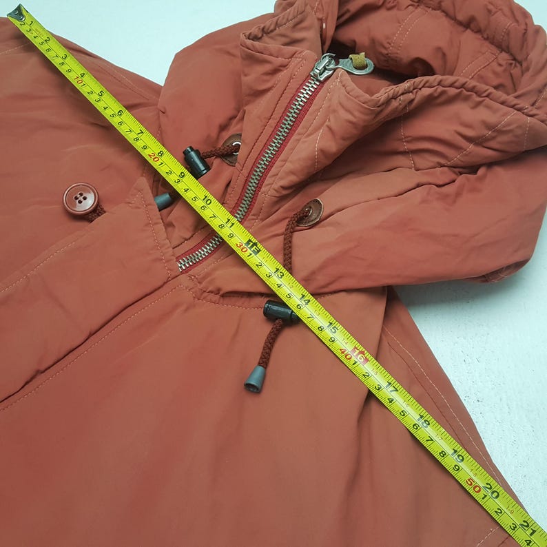 May include: A rust-colored hooded jacket with a zipper and drawstring closure. The jacket features a button detail and a hood. A yellow measuring tape is laid across the jacket, showing a measurement of approximately 21 inches.