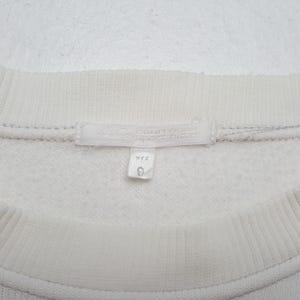 May include: Close-up of a cream-colored sweatshirt. The tag reads "Courreges Sportswear" and includes Japanese text. A small tag below the main tag displays the number "9". The sweatshirt has a ribbed neckline.