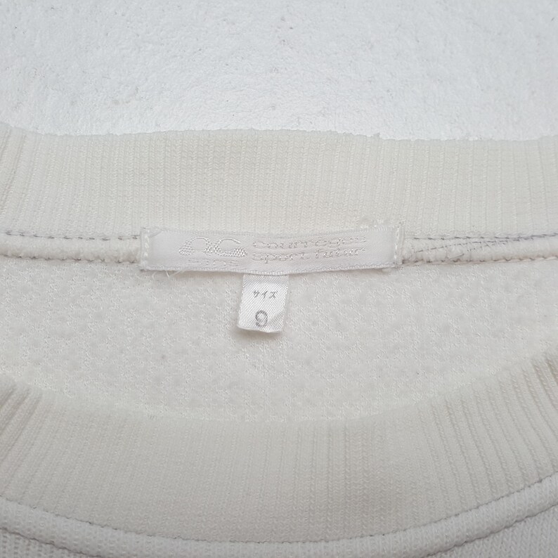 May include: Close-up of a cream-colored sweatshirt. The tag reads "Courreges Sportswear" and includes Japanese text. A small tag below the main tag displays the number "9". The sweatshirt has a ribbed neckline.