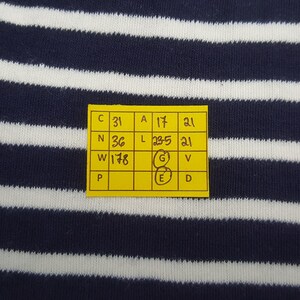May include: Close-up of a navy blue and white striped fabric. A yellow tag with black text and numbers is attached to the fabric. The tag includes measurements and letters. The fabric has horizontal stripes.