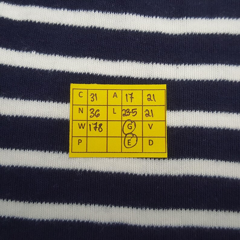 May include: Close-up of a navy blue and white striped fabric. A yellow tag with black text and numbers is attached to the fabric. The tag includes measurements and letters. The fabric has horizontal stripes.