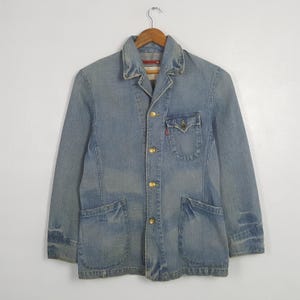 May include: Light blue denim jacket with a classic collar, four gold buttons, and two large front pockets. The jacket has a small pocket on the chest and is hanging on a wooden hanger.
