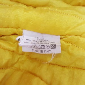 May include: Close-up of a yellow quilted fabric with a white tag. The tag reads "MADE IN ITALY" and lists fabric composition in multiple languages. The fabric has a subtle, textured pattern.