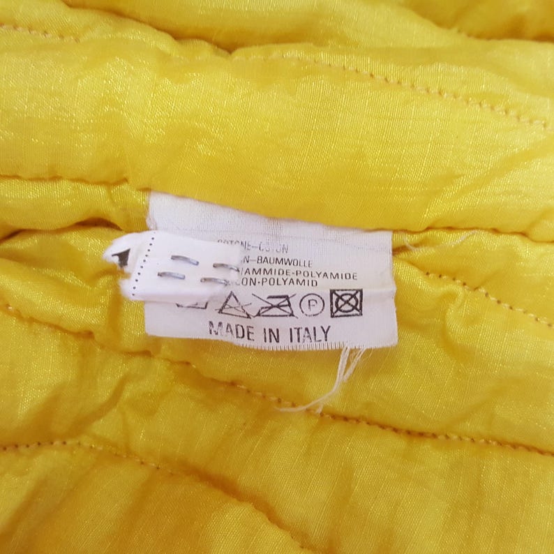 May include: Close-up of a yellow quilted fabric with a white tag. The tag reads "MADE IN ITALY" and lists fabric composition in multiple languages. The fabric has a subtle, textured pattern.