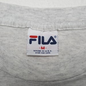 May include: Close-up of a gray t-shirt collar with a Fila brand tag. The tag features the Fila logo in red and navy blue, with the text "-M-" and "MADE IN U.S.A. OVER FOR CARE".