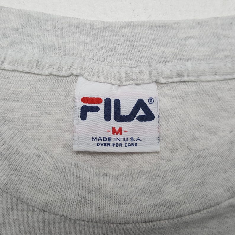 May include: Close-up of a gray t-shirt collar with a Fila brand tag. The tag features the Fila logo in red and navy blue, with the text "-M-" and "MADE IN U.S.A. OVER FOR CARE".