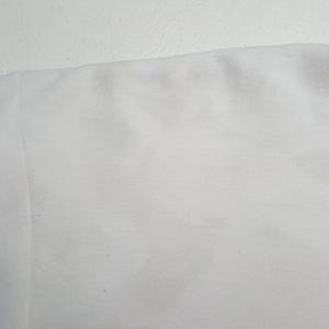 May include: Close-up of a white fabric, possibly a section of a garment. The fabric appears to be a soft, textured material, with subtle variations in tone and slight imperfections.