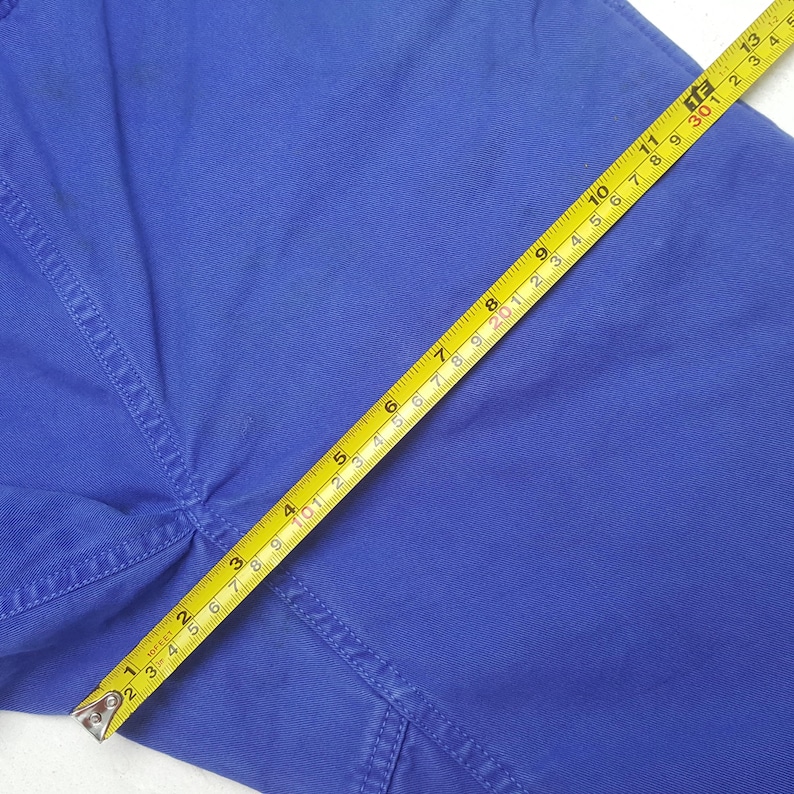 May include: Close-up of blue fabric with a yellow measuring tape. The tape shows measurements in inches, highlighting the texture and color of the material. The fabric appears to be part of a garment.