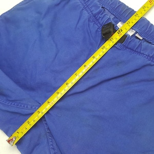 May include: Close-up of blue pants with an elastic waistband. A yellow tape measure shows a measurement of approximately 15 inches. The pants are made of a solid blue fabric.