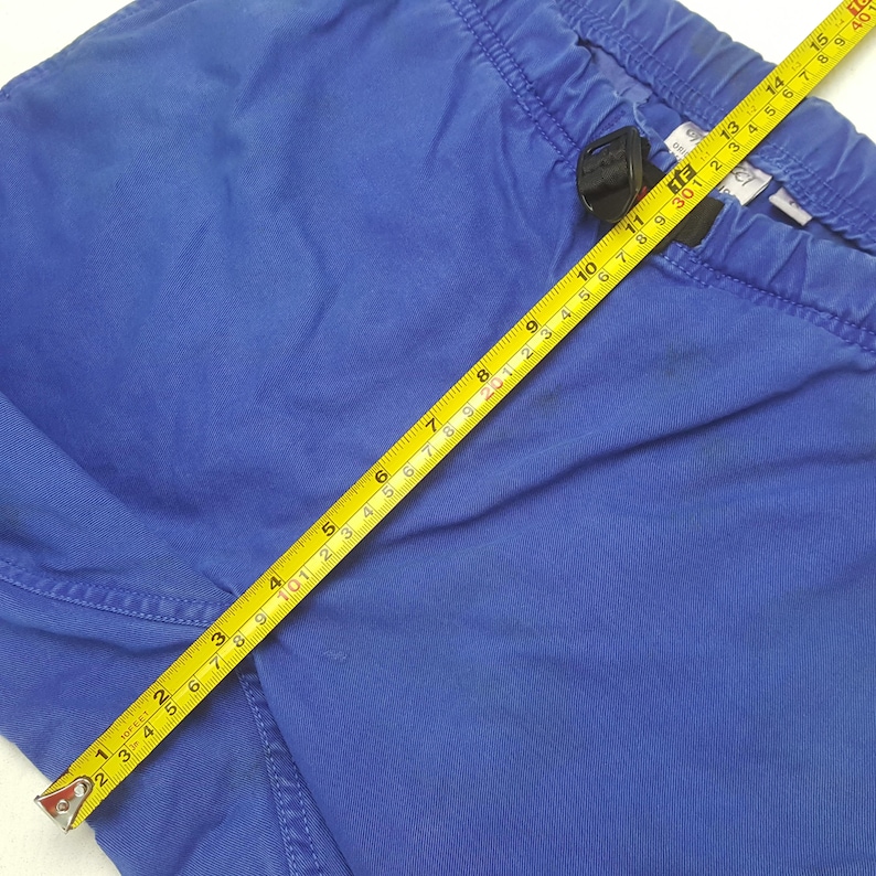 May include: Close-up of blue pants with an elastic waistband. A yellow tape measure shows a measurement of approximately 15 inches. The pants are made of a solid blue fabric.