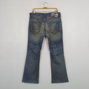 May include: Faded blue denim jeans with a bootcut leg, displayed on a hanger. The jeans feature distressed detailing, a branded patch on the back, and decorative stitching on the back pockets. The jeans are a classic style.