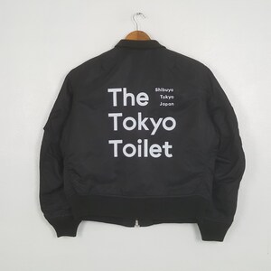 Könnte beinhalten: Schwarze Bomberjacke mit weißem Text "The Tokyo Toilet" und "Shibuya Tokyo Japan" darunter. Die Jacke hängt an einem Kleiderbügel vor weißem Hintergrund.