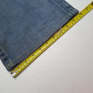 May include: Close-up of a section of blue denim jeans with a yellow measuring tape. The tape shows measurements in inches, highlighting the fabric's texture and the precision of the measurement. The jeans are on a white surface.