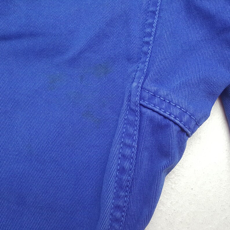 May include: Close-up of a section of blue fabric, possibly denim or a similar material. The fabric has a textured appearance with visible vertical lines and stitching along the edge. There are some darker spots on the fabric.