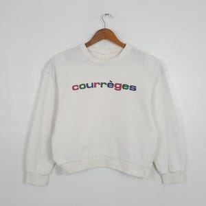 May include: White crew neck sweatshirt with the word "courr&egrave;ges" embroidered across the chest in colorful letters. The sweatshirt has long sleeves and a ribbed hem and cuffs. The garment is hanging on a wooden hanger.