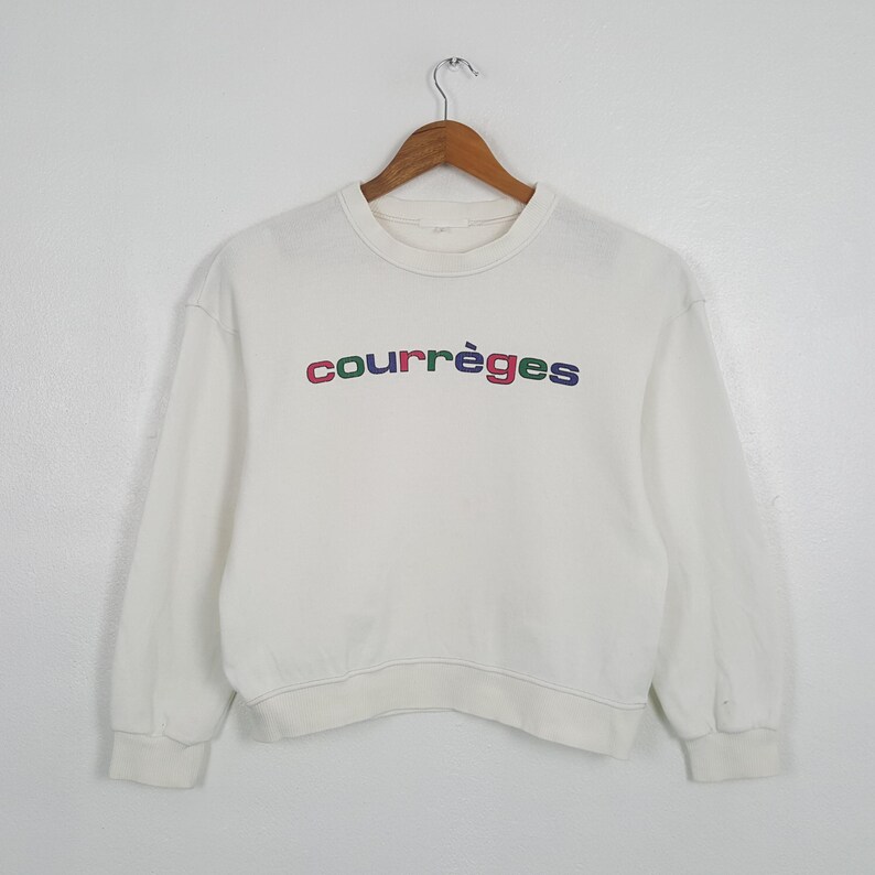 May include: White crew neck sweatshirt with the word "courr&egrave;ges" embroidered across the chest in colorful letters. The sweatshirt has long sleeves and a ribbed hem and cuffs. The garment is hanging on a wooden hanger.
