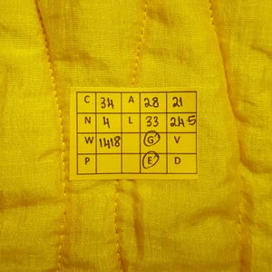 May include: Close-up of a bright yellow fabric with a stitched pattern. A yellow rectangular tag with black text and numbers is attached to the fabric. The tag contains the numbers 34, 28, 21, 4, 33, 245, 1418.