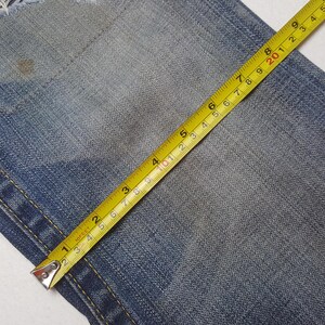 May include: Close-up of blue denim fabric with a yellow measuring tape. The tape shows measurements in inches, with the numbers 1 through 5 clearly visible. The denim has a worn appearance.