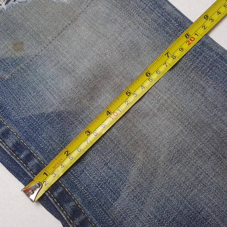 May include: Close-up of blue denim fabric with a yellow measuring tape. The tape shows measurements in inches, with the numbers 1 through 5 clearly visible. The denim has a worn appearance.