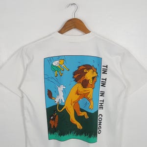 May include: White t-shirt featuring a colorful illustration from the Tintin in the Congo comic. The design includes a roaring lion, a character in green, and the text "TIN TIN IN THE CONGO" in black.